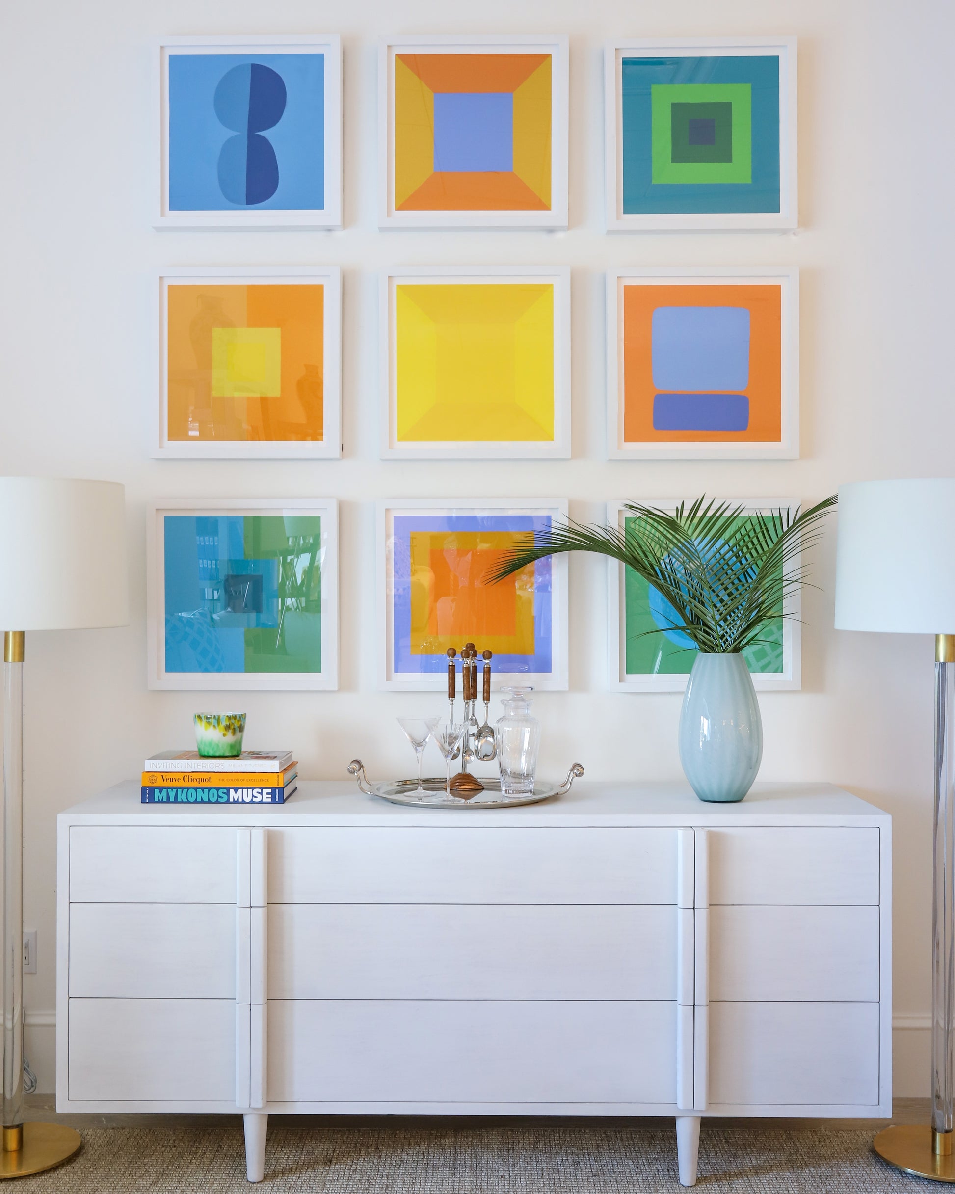 A white sideboard holds a tray, glasses, books, and a palm-leaf vase beneath two lamps and nine Ornis Gallery Modern 6170 geometric abstraction artworks in Gallery Deep White frames on a crisp white wall.