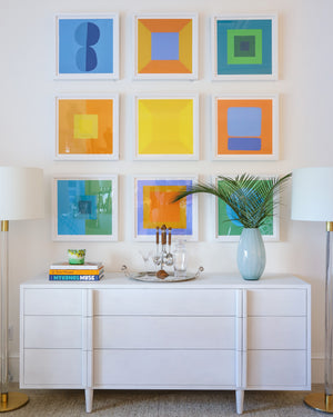 The Modern 6034 by Ornis Gallery features nine geometric abstraction artworks on archival paper and inks, displayed above a white sideboard with books, a glass tray, drinks, and greenery on a pristine white wall.