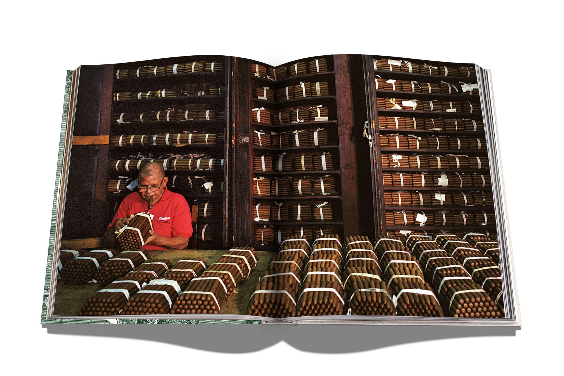 An open book shows a man meticulously arranging exceptional cigars from renowned makers in a room filled with Assouline's "The Impossible Collection of Cigars" on its shelves.