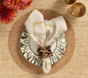 A stylish table setup showcases the Kim Seybert Natural Wood Bead Placemat (15" diameter, cotton backing), a patterned plate, folded napkin in a brown ring, two gold-toned glasses, and orange flowers in the corner.