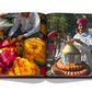A book opened to pages showing people engaging in cultural activities: one side depicts a vendor with colorful flowers, the other a man in traditional attire pouring liquid from a large brass container, all set against the historic backdrop of Jaipur, the enchanting Pink City, is featured in Jaipur Splendor by Assouline.