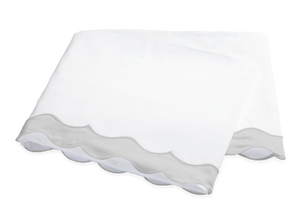 The Lorelei Bedding Collection in Silver by Matouk features a folded white bedsheet with an appliqué scallop edge and light gray border, showcasing modern femininity against a plain background.