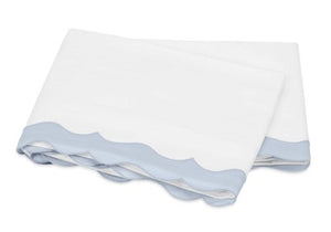 The Matouk Lorelei Bedding Collection in Blue features a folded white sheet with a light blue scalloped trim, made from luxurious 600 thread count Egyptian cotton and OEKO-TEX® certified.