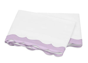 Folded white sheets from the Matouk Lorelei Bedding Collection, Violet, featuring a lavender scalloped edge and crafted from luxurious 600 thread count Egyptian cotton, displayed on a white background.