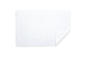 The Matouk Lotus Bath Collection in White features a neatly folded bath mat, crafted from low-twist cotton for extra softness, with a textured border design. Made by Matouk in Portugal, it rests elegantly on a plain background.