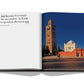 An open Marrakech Flair book by Assouline with a photograph of a historical building in Marrakech and a quote praising the city's vibrant Moroccan arts.