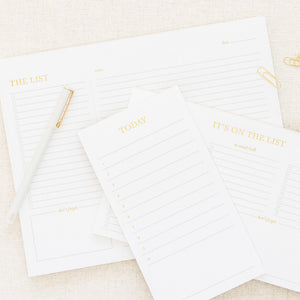 Sugar Paper’s "The List" White Pad set features pads labeled "THE LIST," "TODAY," and "IT'S ON THE LIST," plus a notes section, pen, and paper clips, all arranged on a light fabric surface.
