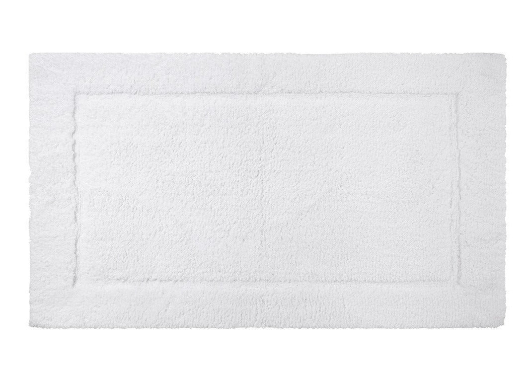The Yves Delorme Prestige Blanc Bath Mat is rectangular, white, and features a simple textured border. Made from 100% cotton, it offers a soft, plush, and luxurious feel.