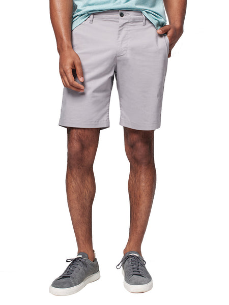 A man wearing Faherty Movement Chino Shorts in light gray and a teal shirt tucked in on one side stands casually with hands in pockets, paired with gray sneakers.