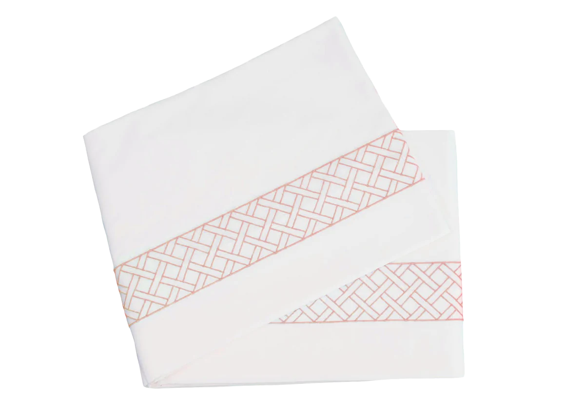 Two folded sheets from the HIVE Exclusive Rovella Bedding Collection in Salmon by Matouk, featuring a pink geometric lattice edge and crafted from Egyptian cotton percale, displayed on a white background.