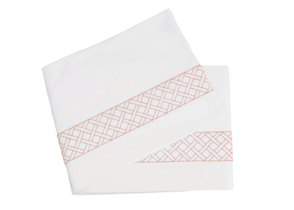 Two folded sheets from the HIVE Exclusive Rovella Bedding Collection in Salmon by Matouk, featuring a pink geometric lattice edge and crafted from Egyptian cotton percale, displayed on a white background.