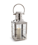 The Nantucket Nickel Outdoor Lantern, Large by Napa Home & Garden features a maritime-inspired silver metal frame, glass panels, a white pillar candle inside, and a round carrying handle for an upscale outdoor look.