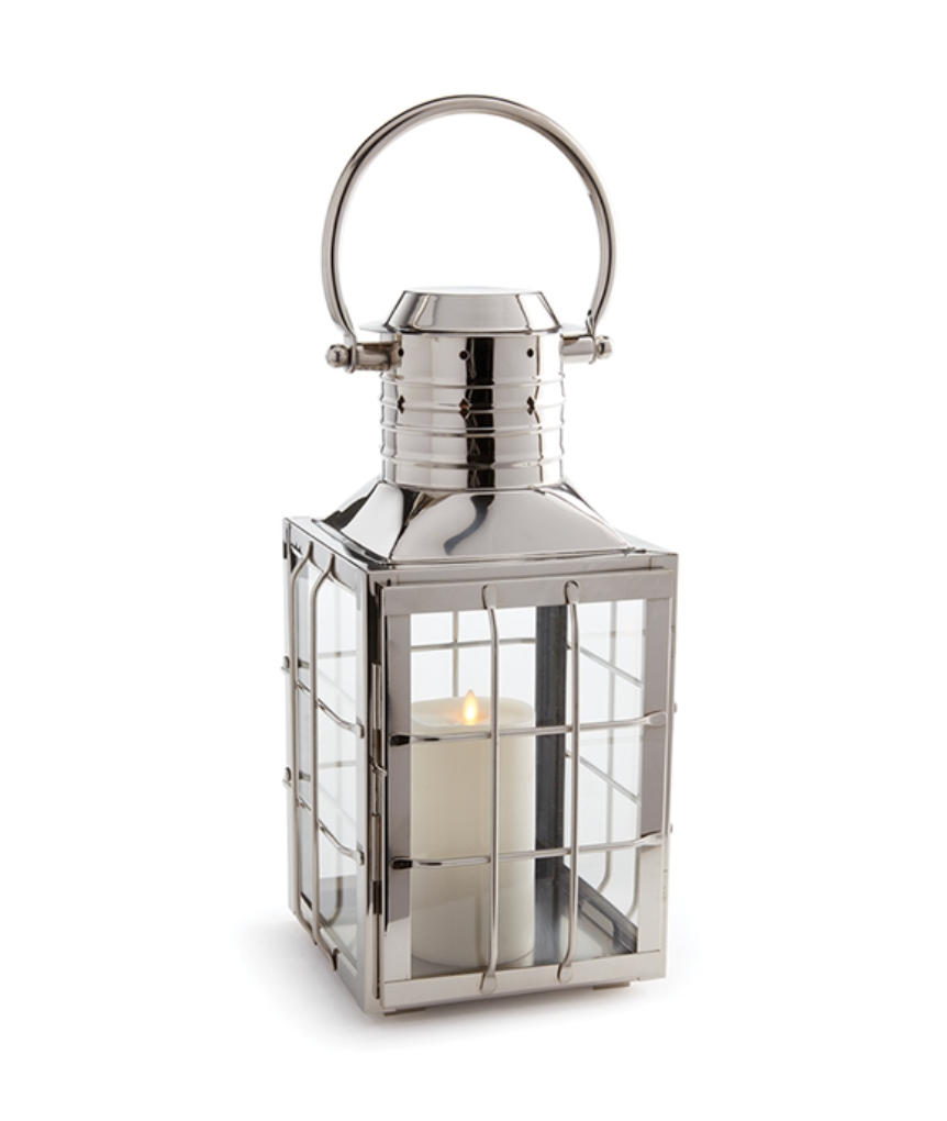 The Nantucket Nickel Outdoor Lantern, Large by Napa Home & Garden features a maritime-inspired silver metal frame, glass panels, a white pillar candle inside, and a round carrying handle for an upscale outdoor look.