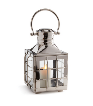 The Napa Home & Garden Nantucket Nickel Outdoor Lantern, Small, features glass sides, a handle, and a lit white candle inside, offering an elegant metallic accent to elevate your outdoor space.