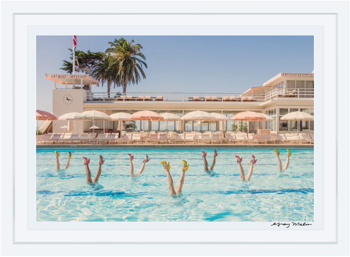 Eight synchronized swimmers with raised legs create a classic Gray Malin vibe, set before a mid-century building and palm trees in "Gray Malin Pool Party, White Frame, 29.5\" x 41.5\"" by Gray Malin—a true poolside scene.