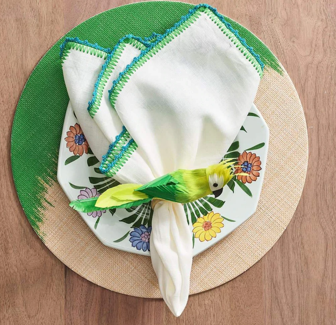 A white napkin with green trim is folded on a floral plate and held by the Kim Seybert Parakeet Napkin Ring, featuring vibrant plumes, set on a green and beige placemat atop a wooden table.