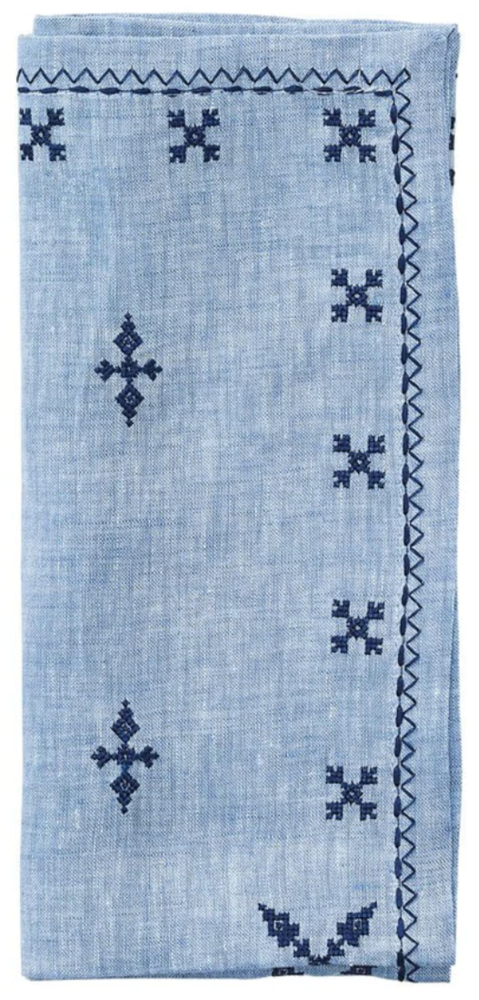 The Kim Seybert Fez Periwinkle Napkin Set features folded light blue linen napkins with navy geometric embroidery and a zigzag stitched border.