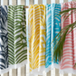 Five striped beach towels, including the Matouk Zebra Palm Beach Towel in Flamingo, in green, navy, yellow, blue, and pink hang side by side on a wooden fence with tropical leaves in the foreground.