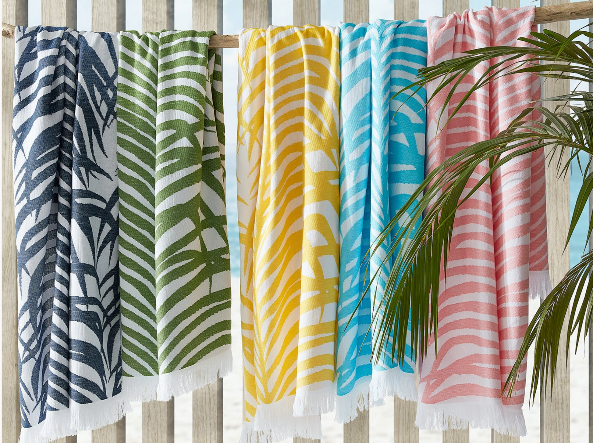 Five striped beach towels, including the Matouk Zebra Palm Beach Towel in Flamingo, in green, navy, yellow, blue, and pink hang side by side on a wooden fence with tropical leaves in the foreground.