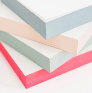A stack of Sugar Paper Painted Pads, featuring vibrant covers in blue, pink, green, and red hues, stands against a white background, perfect for jotting down notes wherever you go.