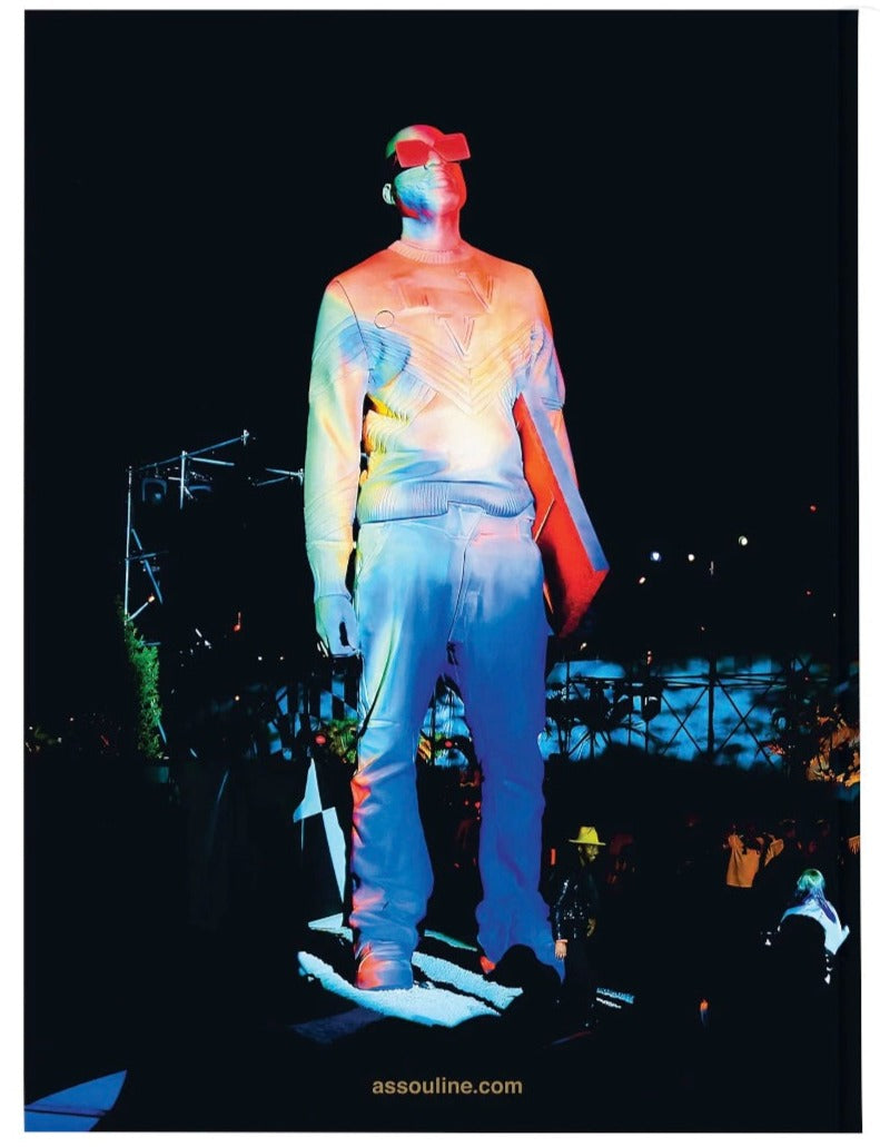 A statue of a person wearing a sweater and pants from the Louis Vuitton: Virgil Abloh by Assouline is displayed outdoors under colorful lighting at night.