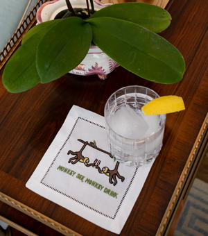 A glass of ice water with lemon rests on an elegant August Morgan Monkey See Monkey Drink cocktail napkin, beside a potted orchid on a wooden table.