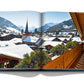An open book displays a scenic winter village with snow-covered rooftops, a church steeple, and mountain peaks in the background, reminiscent of Alpine chic. A wooden balcony railing is visible in the foreground. The book titled "Gstaad Glam" by Assouline captures this perfect winter scene.