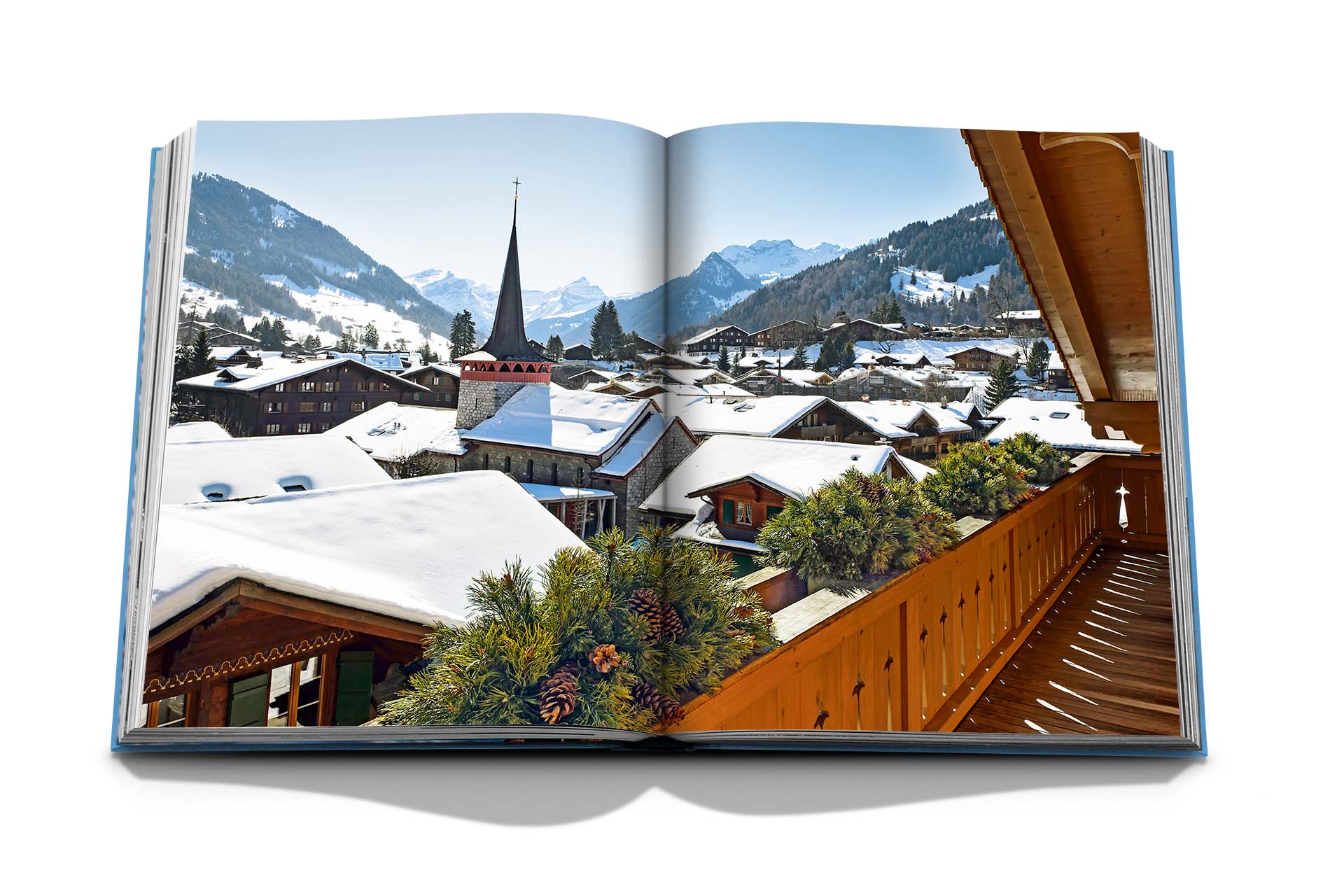 An open book displays a scenic winter village with snow-covered rooftops, a church steeple, and mountain peaks in the background, reminiscent of Alpine chic. A wooden balcony railing is visible in the foreground. The book titled "Gstaad Glam" by Assouline captures this perfect winter scene.
