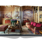 An open book displaying a photo of a cozy living room with wooden walls, red furniture, a fireplace, and an ornate mirror. The room exudes Alpine chic with its classic, rustic design and detailed wall art, reminiscent of the charming chalets in Gstaad in the Swiss Alps. The book is titled "Gstaad Glam" and is published by Assouline.