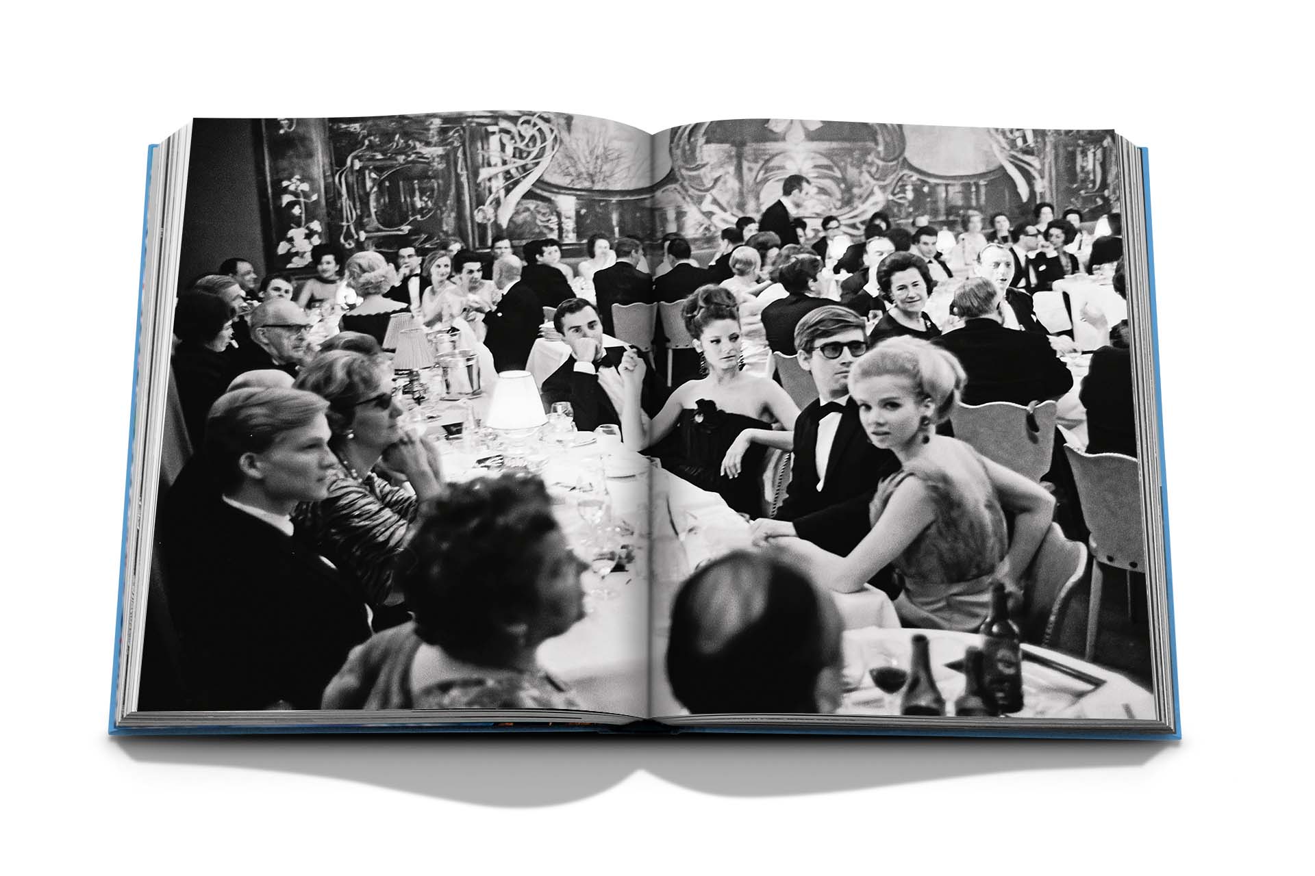 Black and white photo of a formal gathering in an open book. People are seated at round tables, engaging in conversation and dining, with an ornate backdrop reminiscent of Gstaad Glam by Assouline.
