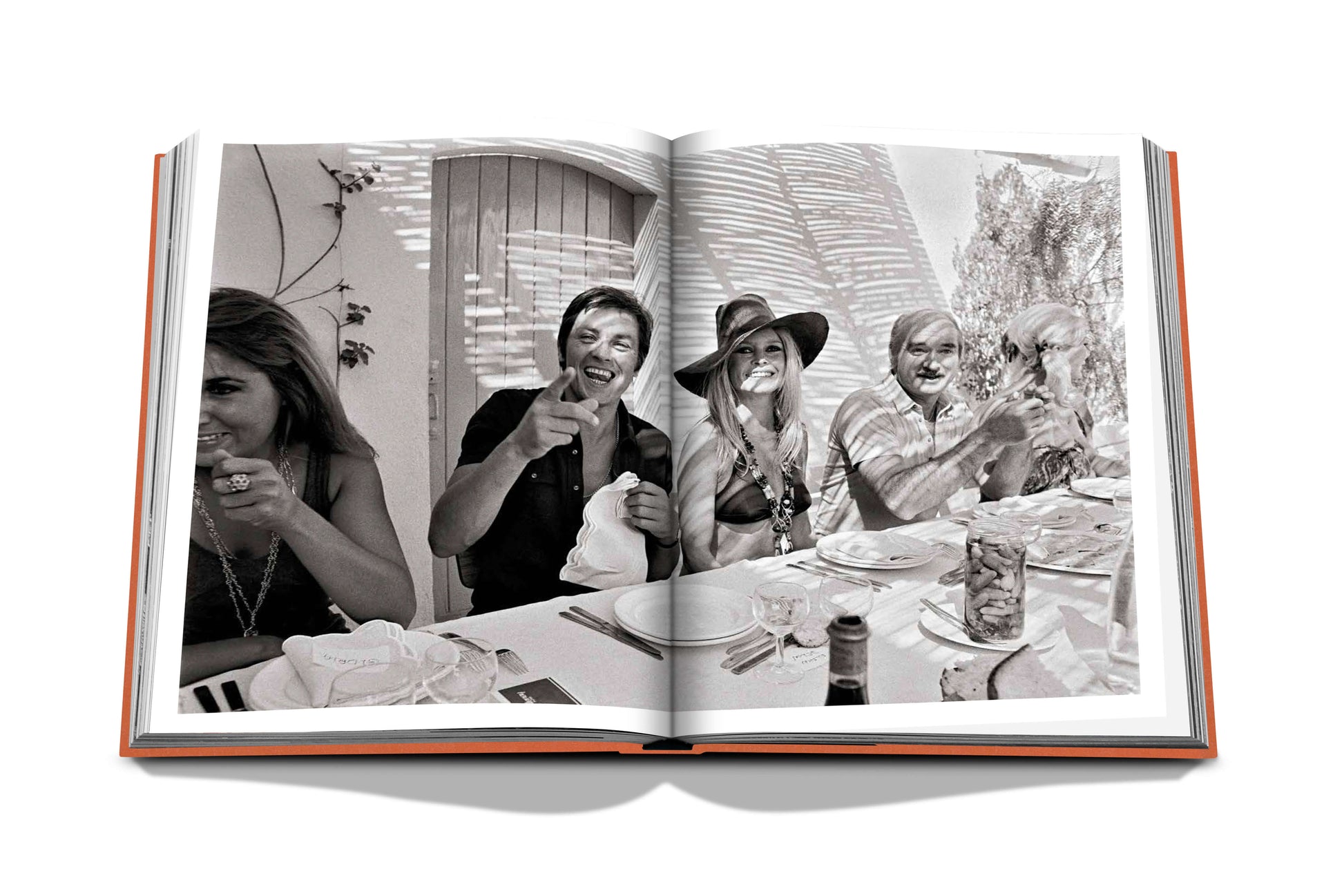 Open book displaying a black-and-white photo of five people enjoying a meal and laughing at an outdoor table, reminiscent of the carefree moments you'd find at places like **St Tropez Soleil by Assouline**.