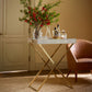 The Aerin Butler Tray Stand with a white tray and solid brass frame displays bottles, decanters, glasses, and a vase of red flowers beside a brown armchair—ideal for stylish entertaining in any sunlit room.