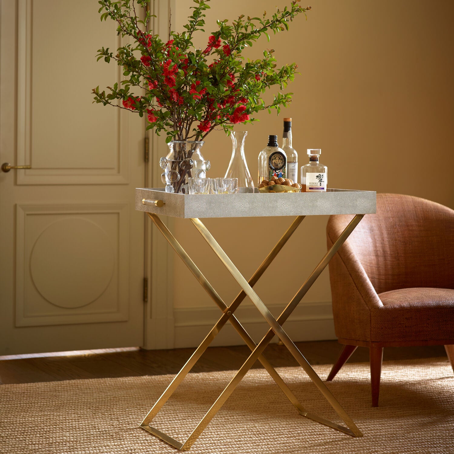 The Aerin Butler Tray Stand with a white tray and solid brass frame displays bottles, decanters, glasses, and a vase of red flowers beside a brown armchair—ideal for stylish entertaining in any sunlit room.