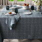 A dining table set with Matouk’s Rubia Navy Napkin Set, plates, apples, glassware, candles, and a vase of white flowers in a room with checkered floor tiles and large windows. OEKO-TEX® Standard 100 certified.