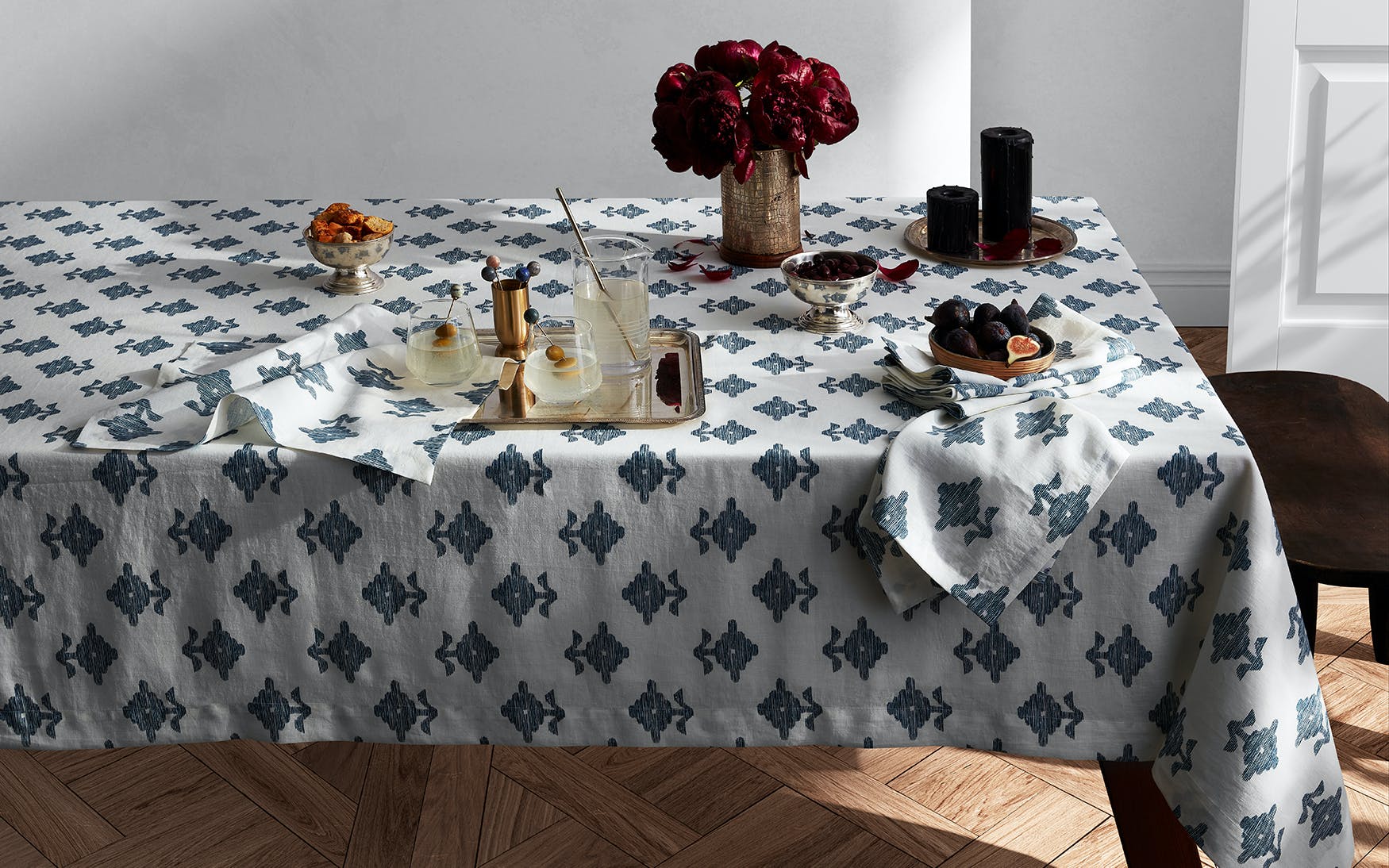 A sunlit dining table with a wooden floor is set with drinks, snacks, fruit, candles, red flowers in a vase, and Matouk Rubia Navy Placemats (set of 4).