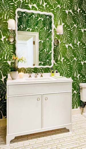 A bathroom featuring green tropical leaf wallpaper, a white vanity, the Carvers' Guild Scalloped Rectangular Mirror, two wall sconces, and a right-side toilet showcases transitional decor for a fresh yet timeless appeal.