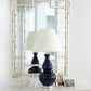 A white dresser with gold handles displays a navy blue lamp, framed photos, and coral decor, set against the Carvers' Guild Bamboo Mirror in Silver featuring an elegant geometric motif.