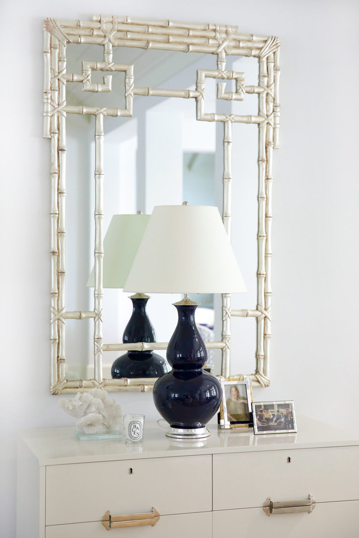 A white dresser with gold handles displays a navy blue lamp, framed photos, and coral decor, set against the Carvers' Guild Bamboo Mirror in Silver featuring an elegant geometric motif.