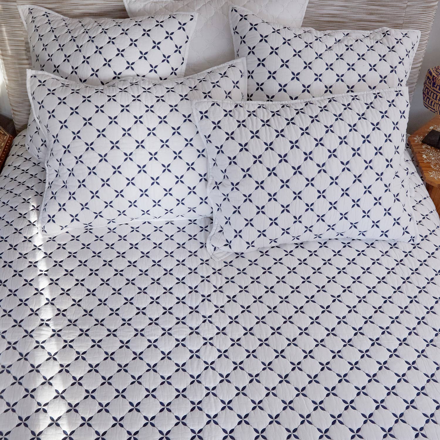 A neatly made bed featuring the John Robshaw Layla Indigo Coverlet and white cotton voile bedding, with pillows adorned in blue geometric patterns inspired by Mughal gardens.