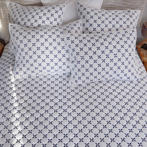 A neatly made bed featuring the John Robshaw Layla Indigo Coverlet and white cotton voile bedding, with pillows adorned in blue geometric patterns inspired by Mughal gardens.