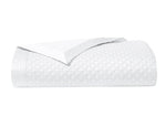 The Adagio Blanc Bedding Collection by Yves Delorme features a neatly folded white quilted blanket with a subtle geometric pattern and smooth satin trim, crafted from luxurious 500 thread count cotton sateen.