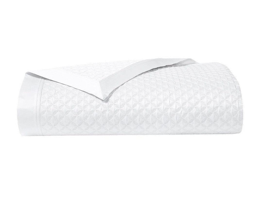 The Adagio Blanc Bedding Collection by Yves Delorme features a neatly folded white quilted blanket with a subtle geometric pattern and smooth satin trim, crafted from luxurious 500 thread count cotton sateen.