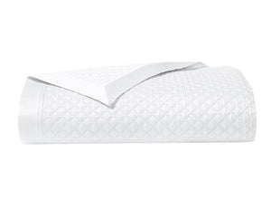 The Adagio Blanc Bedding Collection by Yves Delorme features a neatly folded white quilted blanket with a subtle geometric pattern and smooth satin trim, crafted from luxurious 500 thread count cotton sateen.