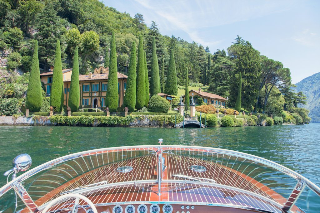 Experience La Dolce Vita with Gray Malin’s “Afternoon Ride Lake Como,” a 24" x 36" framed print capturing a wooden boat gliding toward a villa amid lush gardens and hills, available in a white frame from Gray Malin.