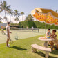 People play tennis on a grass court and sit under a yellow floral umbrella, surrounded by trees and hedges. The scene captures the essence of laid-back elegance on a sunny day in Palm Beach, reminiscent of Gray Malin's luxury prints as featured in "Afternoon Tennis Palm Beach.
