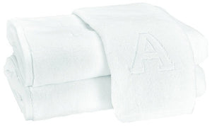 Two Auberge Monogram Bath Towels by Matouk—folded white cotton terry, one with an embroidered "A"—are stacked on a white background, adding elegant charm to any guest bath.