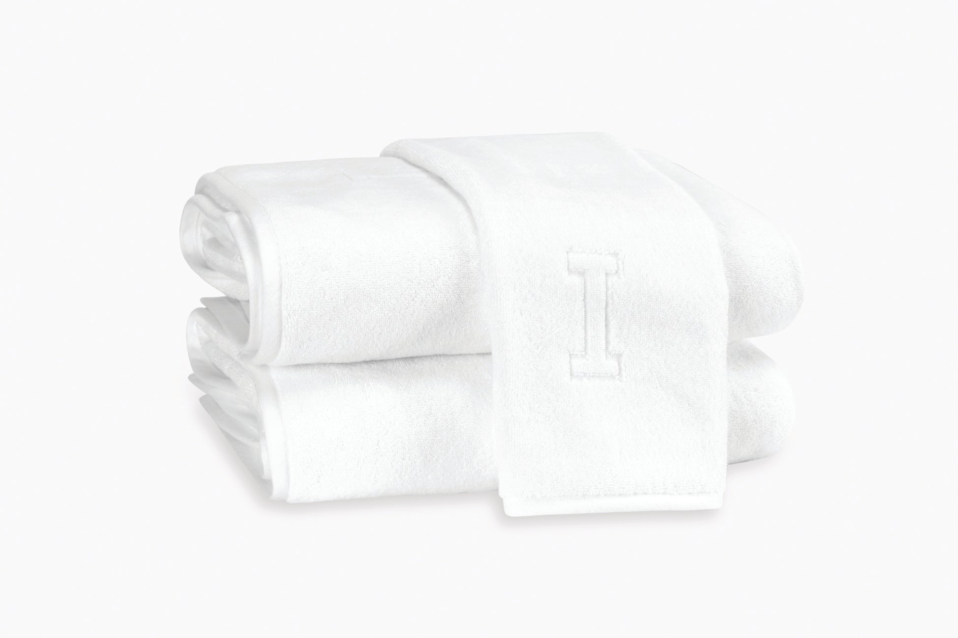 The Matouk Auberge Monogram Bath Towels feature an embroidered “I”; two are folded and stacked with a third draped on top—an elegant addition to any guest bath.