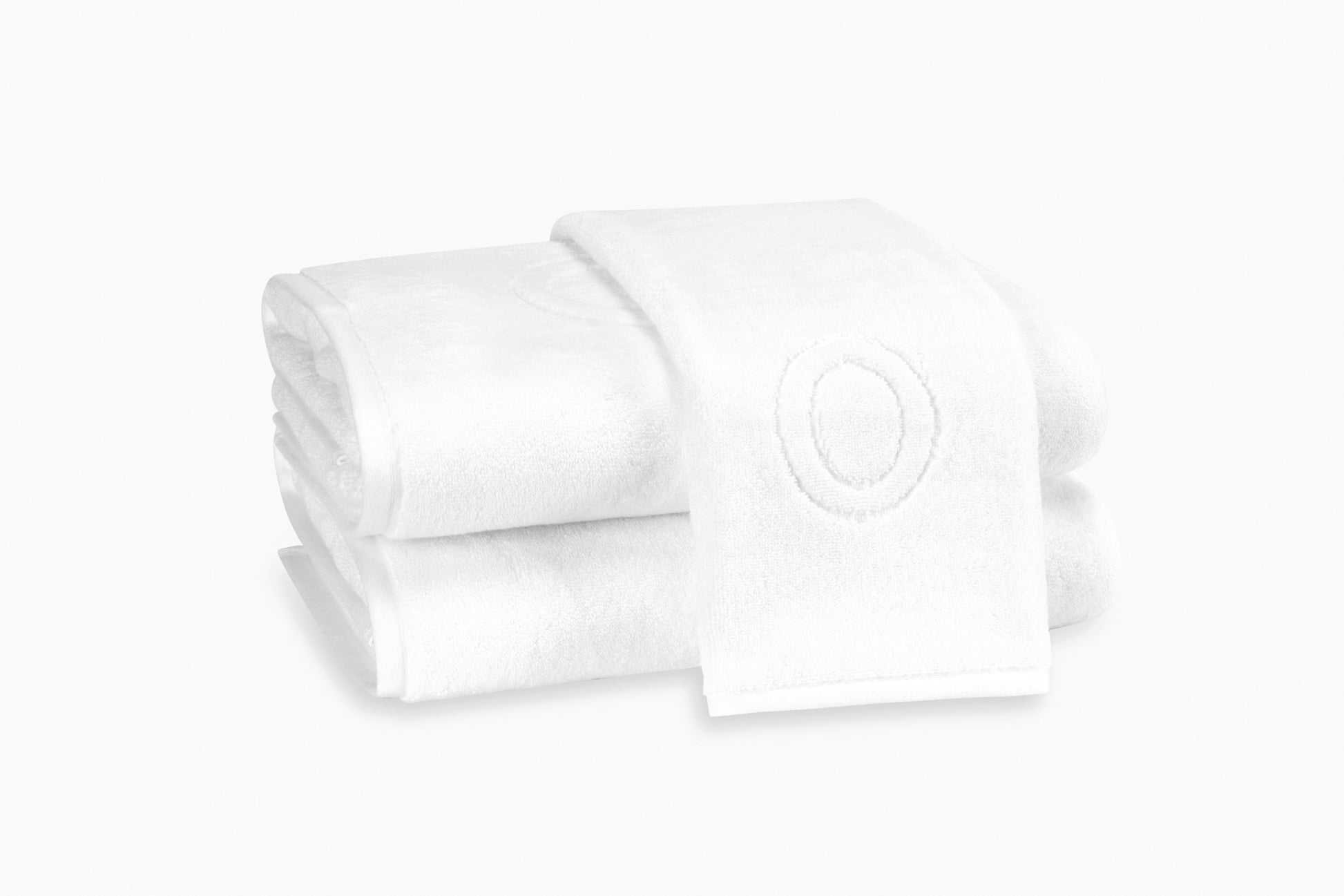 Two neatly folded white Matouk Auberge Monogram Bath Towels are stacked, one displaying an oval monogram design—ideal for elevating any guest bath.