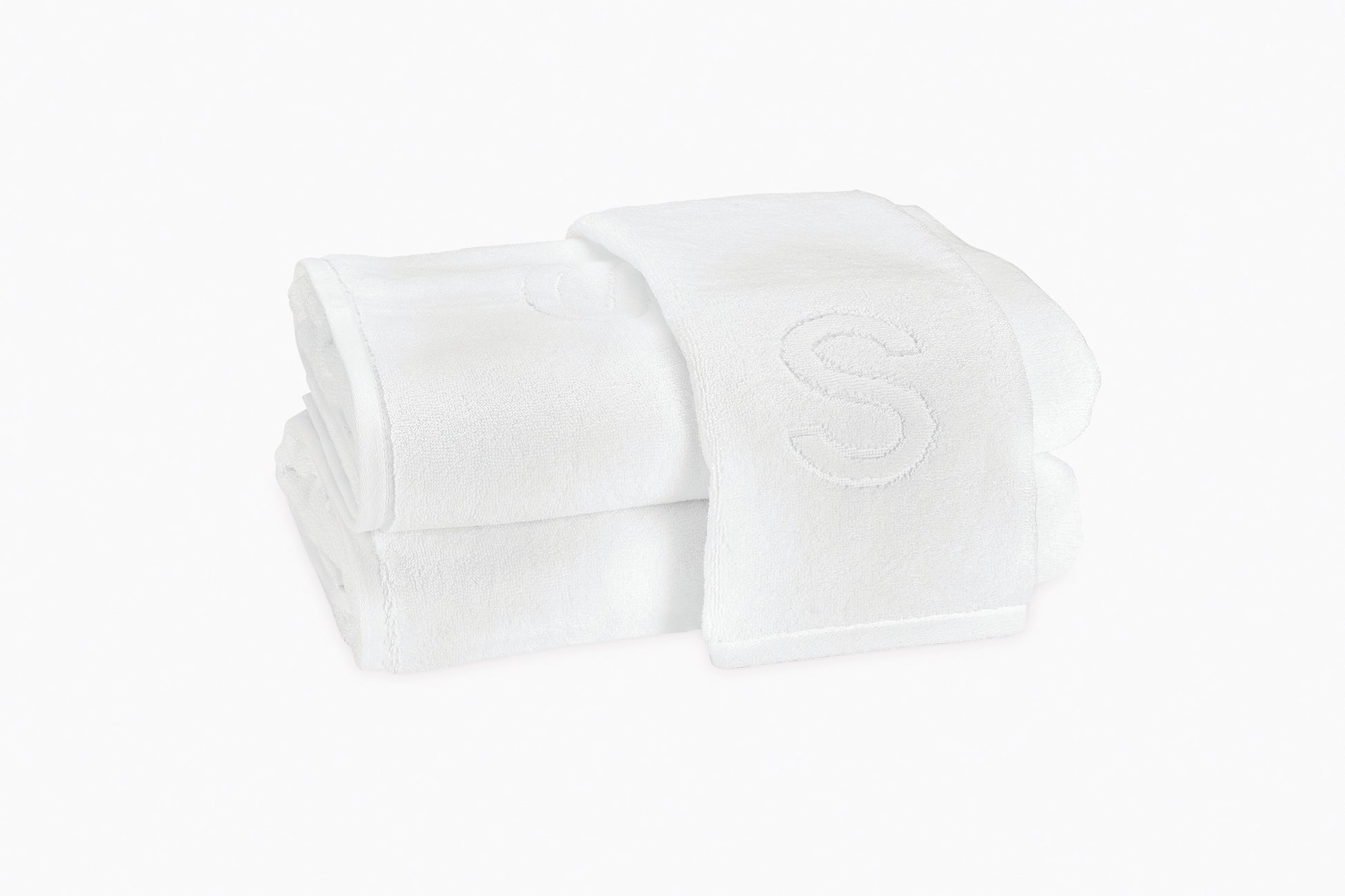Two folded Matouk Auberge Monogram Bath Towels in white cotton terry, with an embroidered "S" on top, bring classic style to any guest bath. Set on a plain white background.