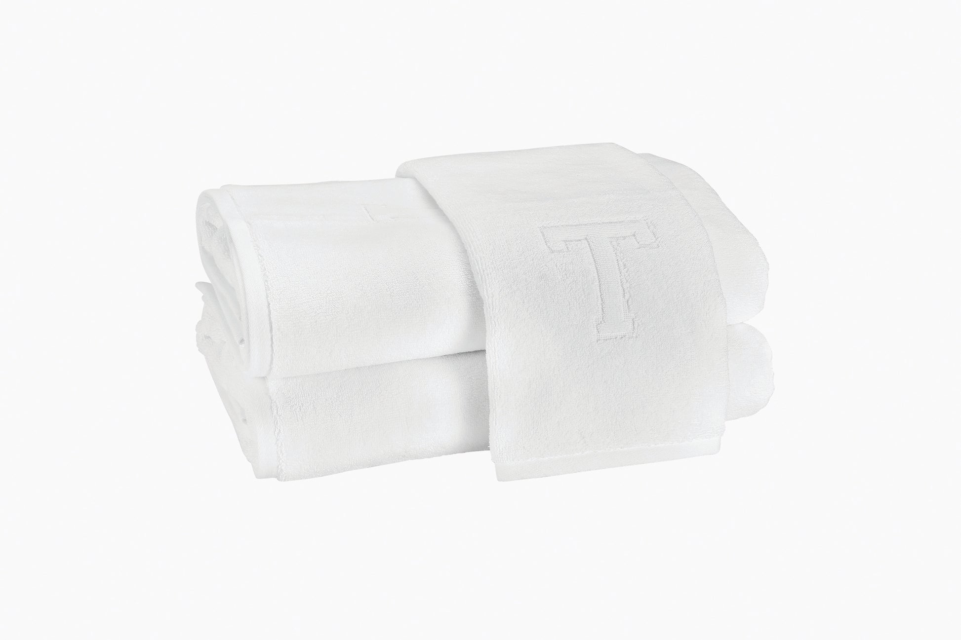 Two folded Auberge Monogram Bath Towels by Matouk, made of white cotton terry with an embroidered "T," ideal for an Auberge-inspired guest bath, shown on a plain white background.